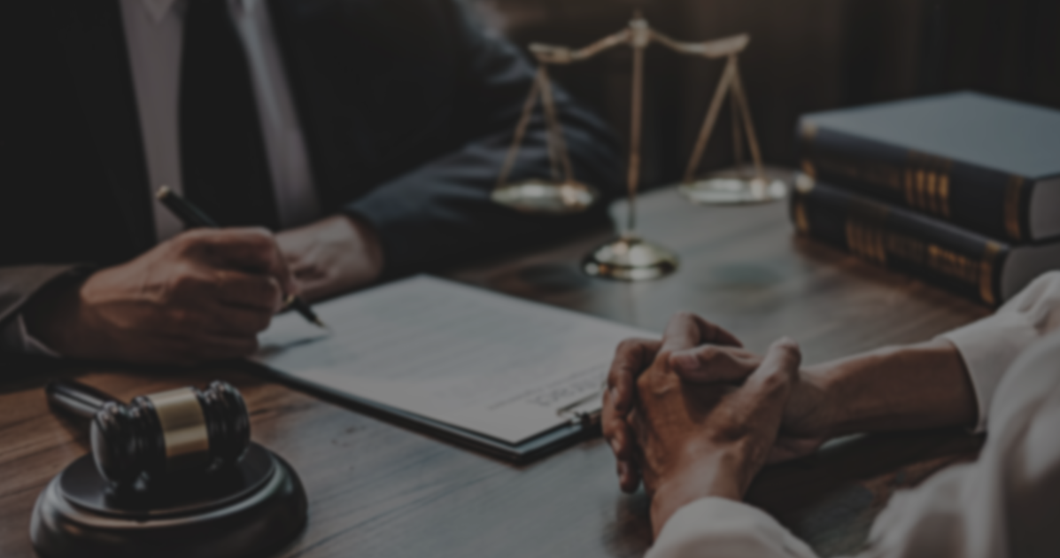 two sets of hands are visible in frame, resting atop a wood table with legal scales on it. on the left, one hand is holding a pen and writing in a clipboard. the other pair is clasped together, as if listening intently.